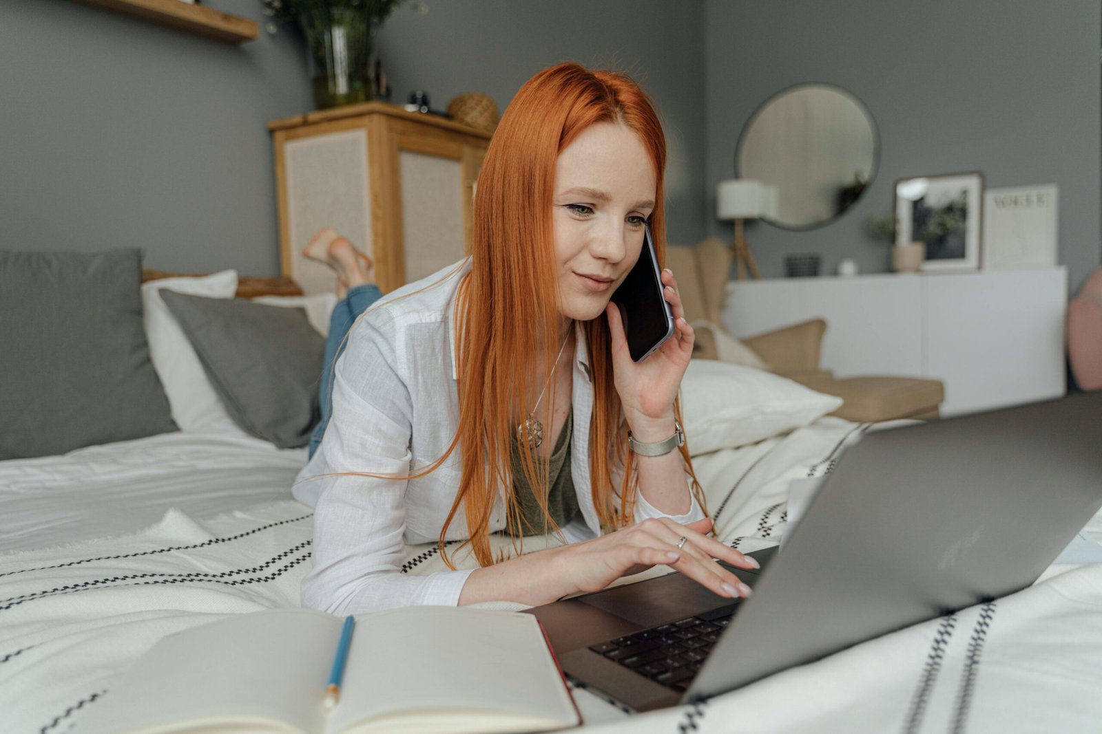 Red-haired woman multitasking in a cozy bedroom, using a laptop and phone for work.