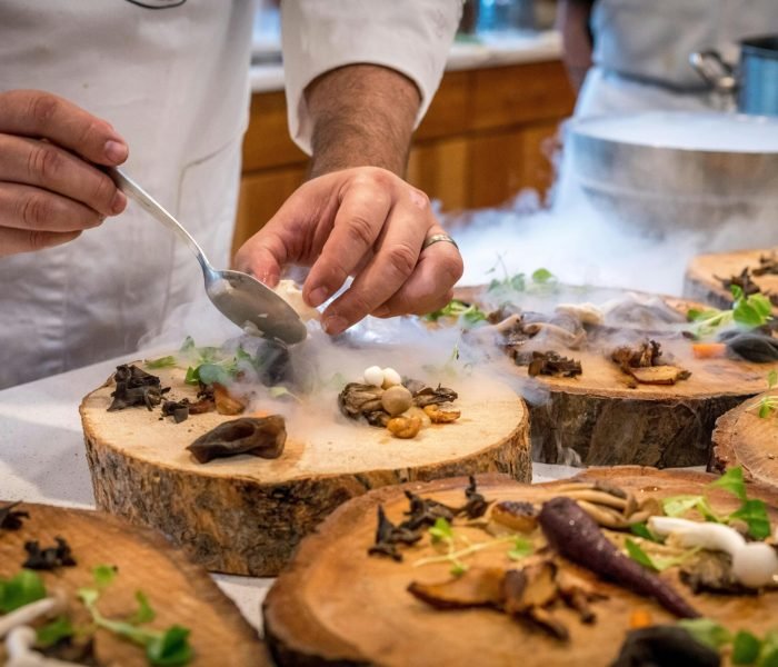 A chef artfully plating a gourmet dish with mushrooms and greens on wood slices.